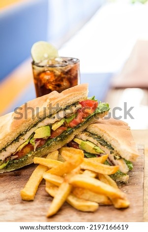 Chicken sandwich, avocado, red onion, tomato, lettuce and pesto, served with french fries and a glass of soda. Served on a wooden board in a restaurant.