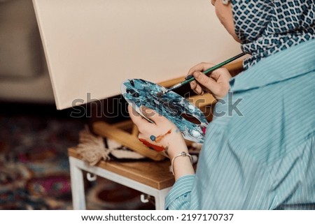 A woman in a hijab paints on canvas with a brush and tempera. 