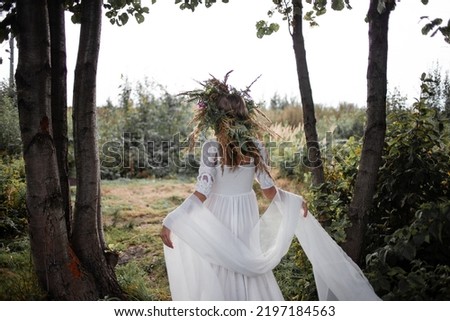 A beautiful young woman in a white sundress and a huge wreath of field herbs poses in a forest clearing, a bride in folk style, back view Royalty-Free Stock Photo #2197184563
