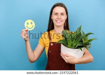 Happy cute young woman gardener holding little funny emoticon and potted plant, smiles broadly wears red uniform, isolated over blue color background wall in studio. Care about house plants concept