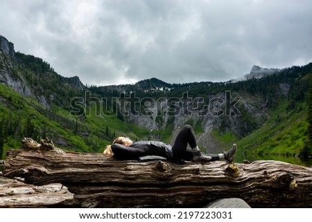 Adventurous athletic female hiker laying down on a log relaxing in front of jagged peaks and an alpine lake in the Pacific Northwest.
 Royalty-Free Stock Photo #2197223031