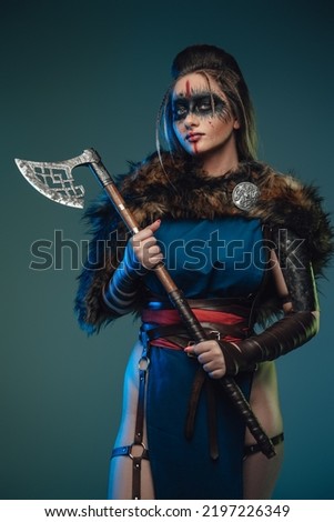Photo of nordic huntress woman with makeup and fur holding axe against teal background.