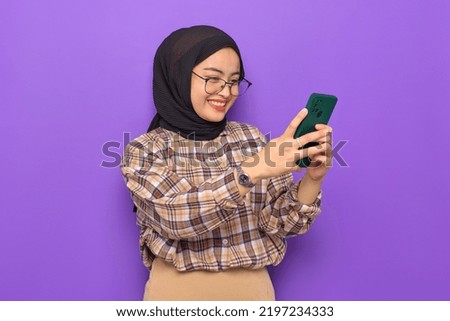 Cheerful young Asian woman in plaid shirt using a mobile phone, getting good news isolated on purple background