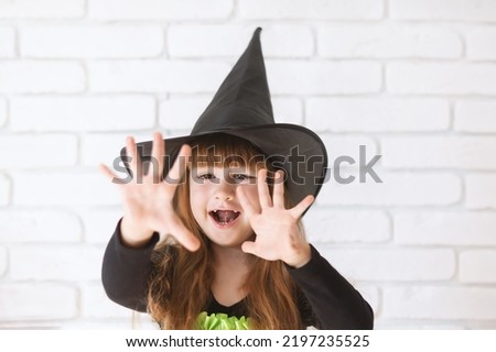 Cute Little Girl In A Witch Hat On A white brick wall Background. Close-up of a 4-year-old girl at the Halloween festival.The little witch in the black hat.Money or life.Traditions, holidays, emotions