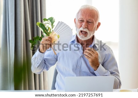 Unwell senior business man sit on desk work on laptop wave with hand fan. Overheated man worker use waver suffer from heatstroke in office, struggle with no AC at workplace.