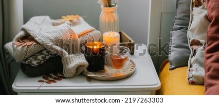 Autumn still life concept. Cozy autumn composition with burning candles, glass cup of tea, stack warm sweaters on bedtable in bedroom. Banner for web site