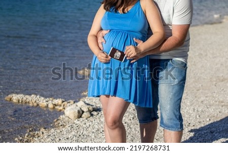 pregnant woman and man husband lover on pebbles lake beach hug holding with hands the big advanced belly.couple with 3d scan ultrasound picture of unborn yet baby.awaiting expecting pregnancy period 