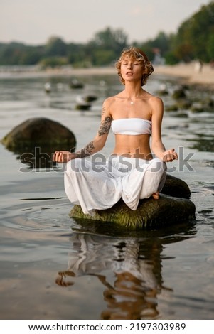 peaceful woman practices yoga position doing meditation in tranquil outdoor sitting on stone in water. Mental wellbeing healthy lifestyle. Royalty-Free Stock Photo #2197303989