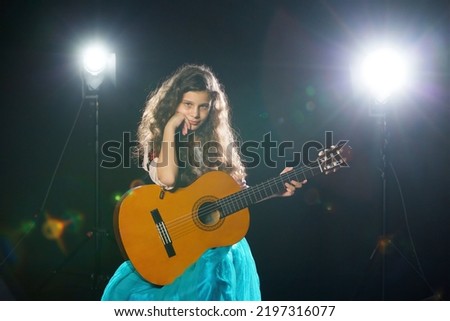 Charming little long-haired girl in a blue dress performs at a solo concert with an acoustic guitar under a bright light