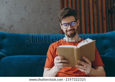 Young smiling man 20s wears red t-shirt glasses read book novel sit on blue sofa couch stay at home hotel flat rest relax spend free spare time in living room indoors grey wall. People lounge concept Royalty-Free Stock Photo #2197322325