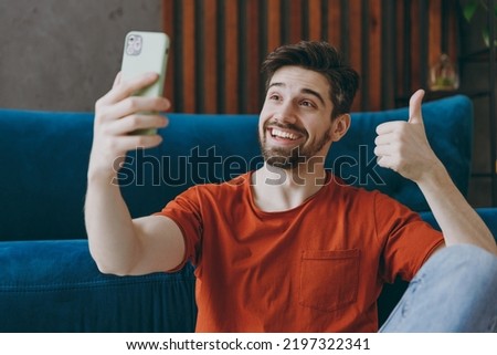 Young man wear red t-shirt do selfie shot on mobile cell phone post photo on social network show thumb up sit on blue sofa coch stay at home flat rest relax spend free spare time in living room indoor