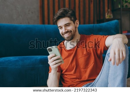 Young man wears red t-shirt hold in hand use mobile cell phone sit on blue sofa couch stay at home hotel flat rest relax spend free spare time in living room indoors grey wall. People lounge concept Royalty-Free Stock Photo #2197322343