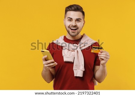 Young excited amazed man in red t-shirt using mobile cell phone hold credit bank card doing online shopping order delivery booking tour isolated on plain yellow color wall background studio portrait