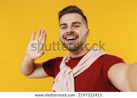 Close up young friendly cheerful cool happy smiling caucasian man 20s wearing red t-shirt casual clothes waving hand say hello greeting isolated on plain yellow color wall background studio portrait.