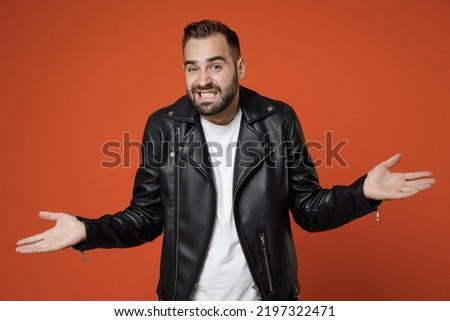 Confused puzzled dissatisfied young bearded man wearing basic white t-shirt black leather jacket standing spreading hands looking camera isolated on bright orange colour background studio portrait Royalty-Free Stock Photo #2197322471