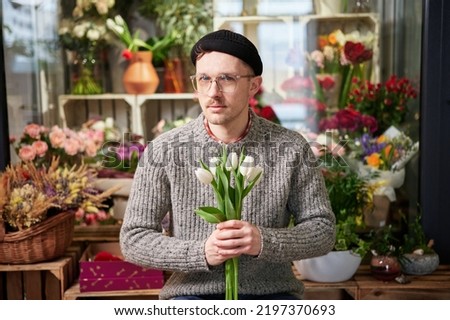 Attractive young caucasian man wearing eyeglasses and beanie in flower store with tulips bouquet. Good looking male florist in sweater holding bouquet. Small business concept. High quality image
