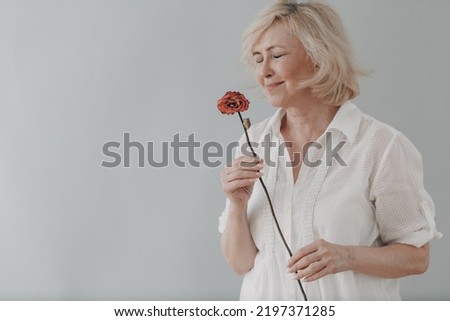 Sad elderly senior woman in white shirt holds withered one dry old dead rose flower. Old age and aging concept.