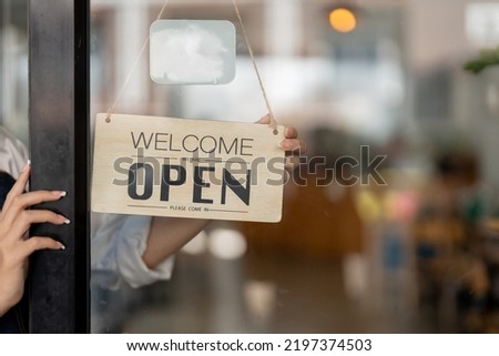 Small business owner smiling while turning the sign for the opening of the place after the quarantine due to covid-19. Close up of woman hands holding sign now we are open support local business.