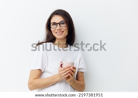 a sweet, beautiful, pleasant woman in glasses and a white t-shirt stands holding a cup in her hands and smiles sweetly.Horizontal photo on a light background
