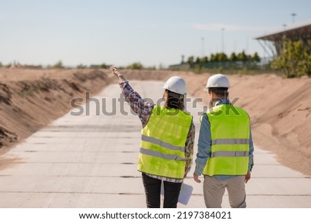 two engineers, a man and a woman in white helmets and protective vests, are standing on the street and holding a plan of the construction and fire safety facility in their hands. engineers designers Royalty-Free Stock Photo #2197384021