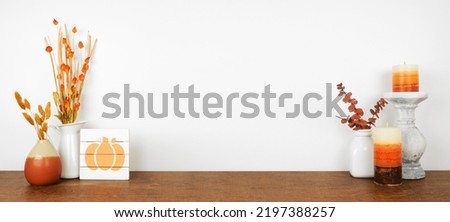 Autumn decor on a wood shelf against a white wall banner background. Pumpkin sign, candles and vases with fall colors. 