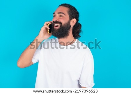 Happy joyful charming young bearded man wearing white T-shirt over blue background talk speak phone smile good mood