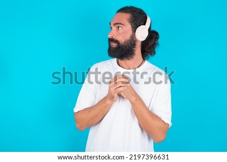 young bearded man wearing white T-shirt over blue background wears stereo headphones listening to music concentrated and looking aside with interest.