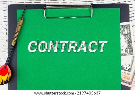 CONTRACT - word (text), a dart for darts and dollar bills on a green background of a notebook, a wooden white table. Business concept: buying, selling, commerce (copy space).