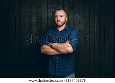 Portrait of a bearded man with a serious look, standing on a black wooden background. a business man in a blue shirt and jeans Royalty-Free Stock Photo #2197408879