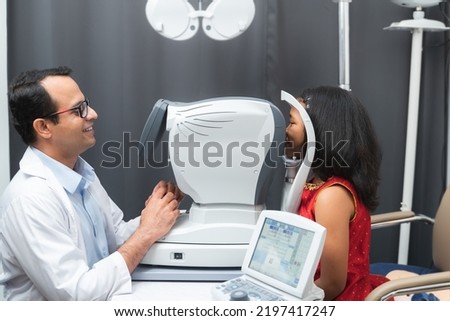 Indian kind man doctor or optometrist in white coat with eyeglasses using Auto refractometer to examine little Indian girl patient's eyesight at hospital or optometry clinic