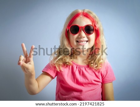 Cute little girl wearing red sunglasses and showing peace sign on gray background.