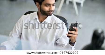Busy indian young business man corporate manager, professional employee, ceo executive holding smartphone using cell phone mobile applications checking financial market data at work in office.