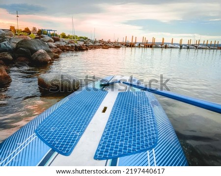 A Stand-Up Paddle Board and Oar Resting on the Shore at dawn. Located within a bay of water with views of a pier with moored sailboats and the coastline. Dawn or dusk dramatic landscape scene.