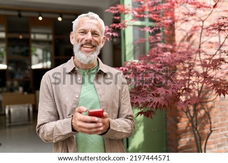 Smiling gray-haired older middle aged bearded man using mobile phone outdoors. Happy old senior adult male user holding cellphone texting on smartphone looking at camera standing outside. Royalty-Free Stock Photo #2197444571