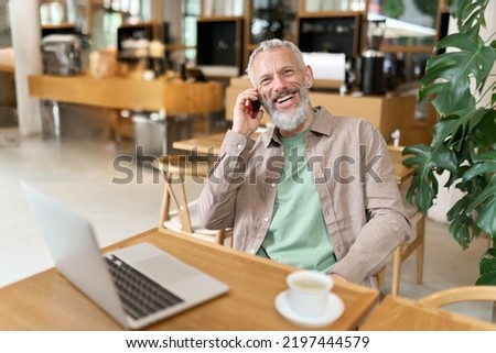 Happy smiling middle aged professional business man, older mature entrepreneur sitting at cafe table talking on cell phone making mobile call remote working online on laptop and having morning coffee. Royalty-Free Stock Photo #2197444579