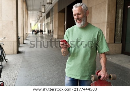 Happy mature older middle aged bearded man using mobile cell phone standing outside. Smiling old adult male hipster with skateboard holding cellphone texting message on smartphone digital technology. Royalty-Free Stock Photo #2197444583