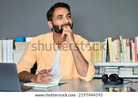 Thoughtful dreamy indian business man writer or student working or learning online, listening podcast looking away thinking of new ideas sitting in office at home using computer, planning project.