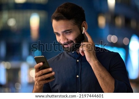 Smiling bearded indian man wearing earbud holding phone having video call at night. Eastern businessman in earphone using smartphone listening music in app tech on cellphone watching videos online. Royalty-Free Stock Photo #2197444601