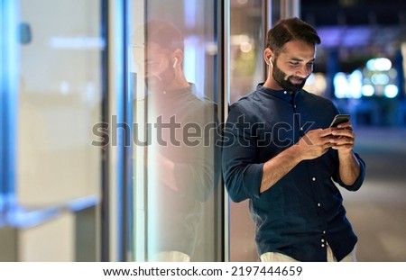 Smiling indian man wearing earbud holding mobile phone listening music at night outdoors, using smartphone having chat call in app tech on cellphone, watching streaming tv videos online. Royalty-Free Stock Photo #2197444659