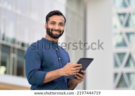 Smiling young adult indian business man professional executive holding digital tablet online fintech outside, happy eastern businessman ceo using pad standing outdoors in big urban city street. Royalty-Free Stock Photo #2197444719