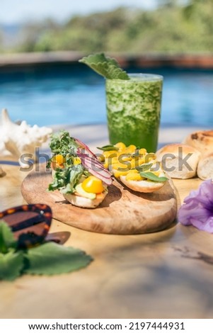 Vegetarian sandwich with white bread, tomato, lettuce, egg and mustard. Served on a wooden plate. Green juice served in a glass of glass. Bread, sunglasses and flowers in the scene. Healthy food