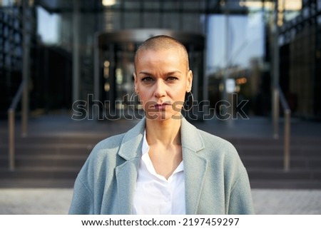 Serious middle-aged empowered woman with shaved hair looking at camera. Business people outdoors on background with buildings corporate. Royalty-Free Stock Photo #2197459297