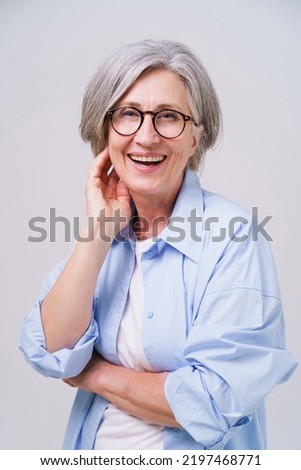 Cheerful pretty grey haired mature business woman in blue shirt and eye glasses stands with hands folded looking straight in camera isolated on white background. Mature people beauty concept.