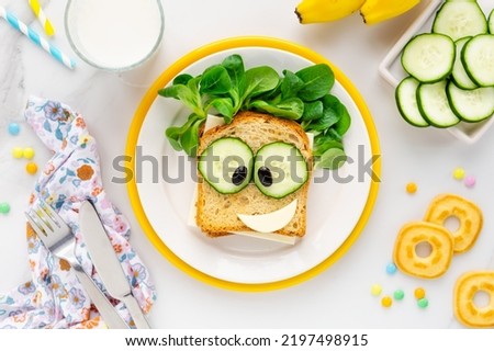 Fun Food for kids - cute smiling cartoon face sandwich with cheese, fresh cucumbers and green corn salad on a yellow palte served with milk and biscuits for breakfast or lunch