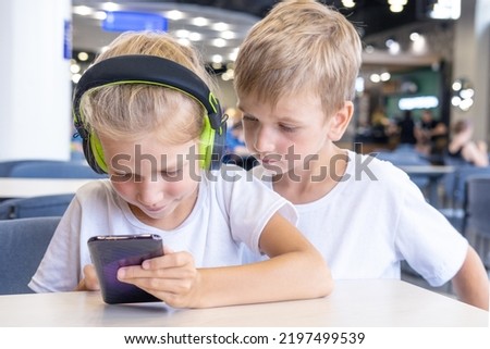 A little girl with headphones and a boy are watching cartoons on their phone, sitting at a table in a cafe. Children listen to music using a mobile phone and headphones. Education, lifestyle
