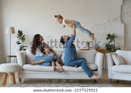 Couple with kids gathered in living room play together have fun sit on sofa, loving dad lifts on arms cute daughter, mom with son embracing resting on couch. Well-being family weekend at home concept
