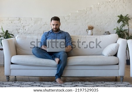 Handsome serious man sits on sofa with computer on laps, spend time at home web surfing information, use modern tech, make order on internet. Freelancing, telework, new software learning, tech concept Royalty-Free Stock Photo #2197529035