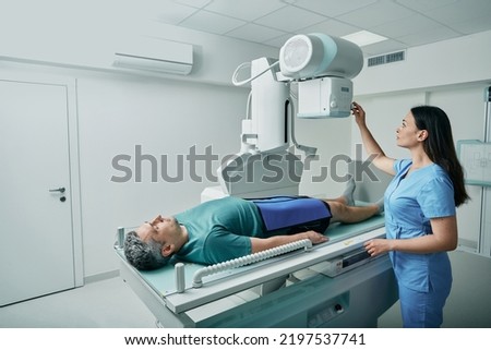 Male patient lying on bed while female nurse adjusting modern X-ray machine for scanning his leg or knee for injuries and fractures Royalty-Free Stock Photo #2197537741