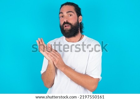 Surprised emotional young bearded man wearing white T-shirt over blue studio background rubs palms and stares at camera with disbelief