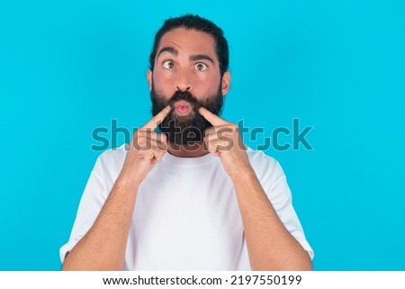 young bearded man wearing white T-shirt over blue studio background crosses eyes and makes fish lips funny grimace